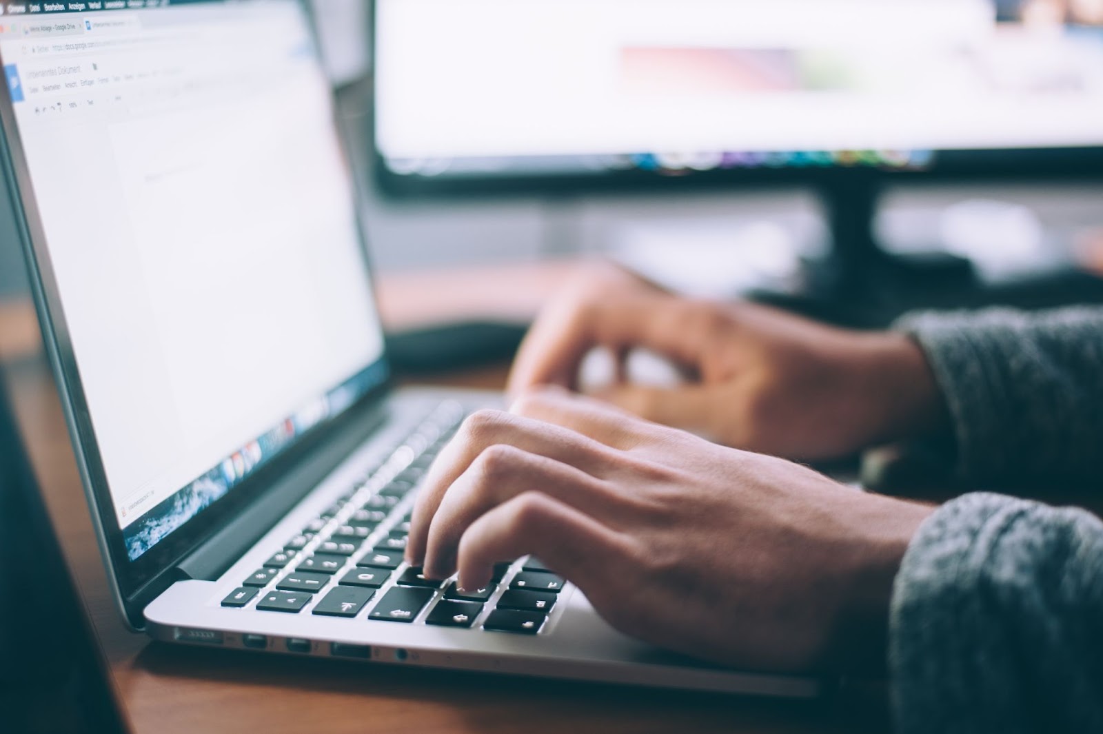 A set of hands typing on a laptop computer in a dim room with a computer monitor in the background.